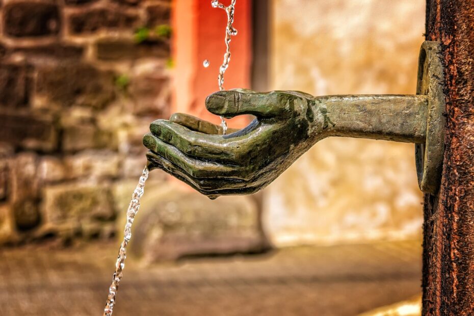 Titel des Bildes: Schön Bibelverse Ermutigung - Das Bild zeigt eine kunstvolle, bronzene Wasserquelle in Form einer ausgestreckten Hand, die Wasser in die Luft sprudeln lässt. Die filigrane Gestaltung der Hand verleiht dem Brunnen eine besondere Symbolik: Sie erinnert an Großzügigkeit, Segen und das Geben und Empfangen von Gnade. Ein schöner Bibelvers zur Ermutigung, der gut zu diesem Bild passt, ist: "Wer aber von dem Wasser trinkt, das ich ihm geben werde, den wird in Ewigkeit nicht dürsten; sondern das Wasser, das ich ihm geben werde, wird in ihm eine Quelle des Wassers werden, das in das ewige Leben quillt." (Johannes 4,14) Dieser Vers erinnert uns daran, dass Gottes Gnade wie lebendiges Wasser ist – erfrischend, belebend und ewig sättigend. Die ausgestreckte Hand im Bild könnte für Gottes Hand stehen, die seinen Segen und seine Liebe über uns ausgießt. Es ist eine wunderbare Erinnerung daran, dass wir durch den Glauben immer wieder neue Kraft und Hoffnung schöpfen können.